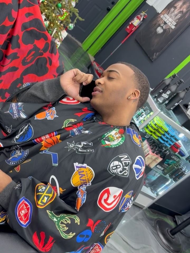 Client on the phone in the chair, fresh fade and line up, wearing a cape covered in team logos